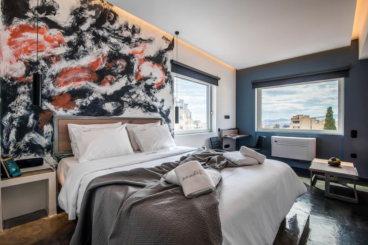 A modern hotel room with a large abstract painting on the wall, a bed with white linens, a bedside table with a book, and a view of the city through large windows.