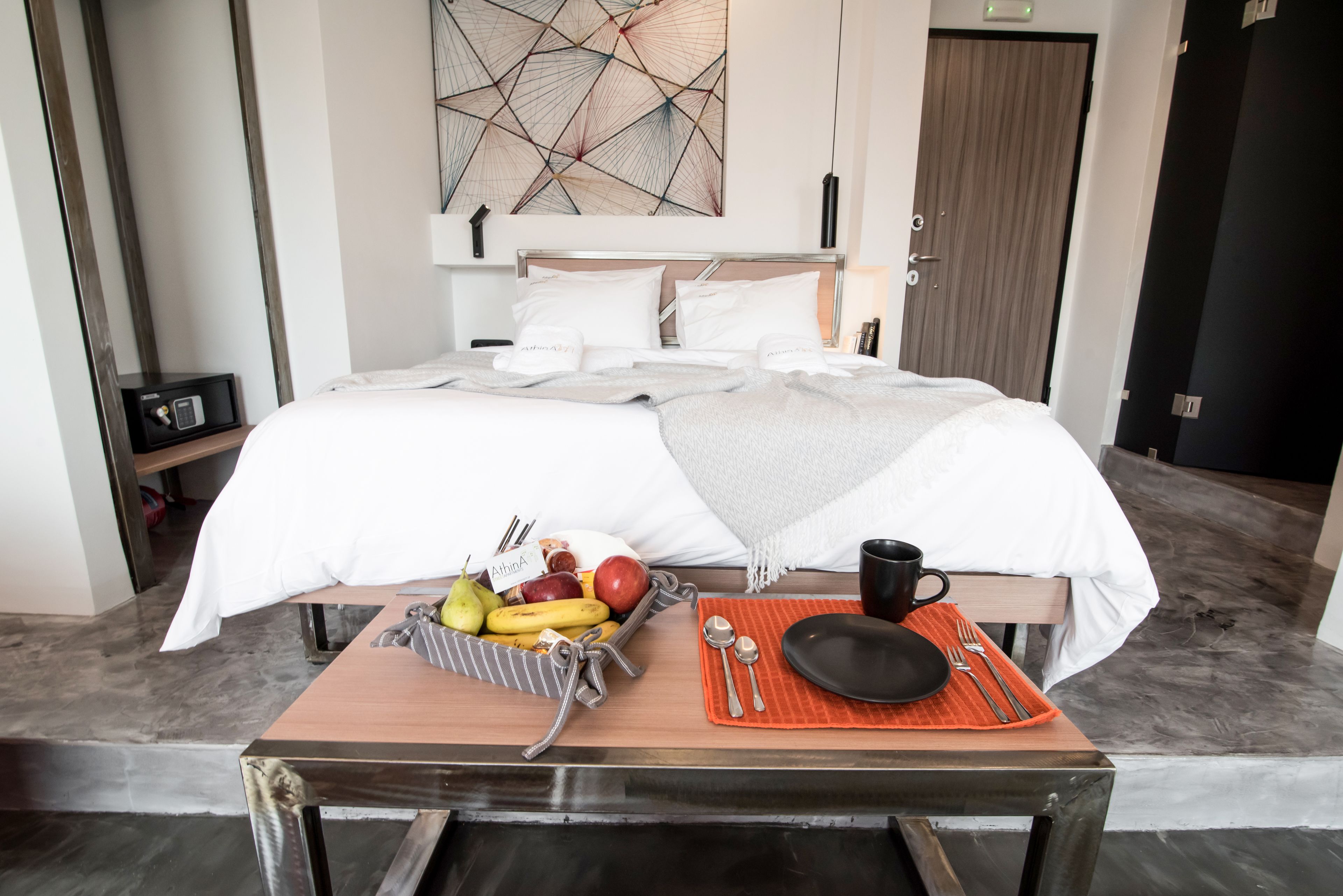 Una camera d'albergo con un letto rifatto, un vassoio per la colazione con frutta e una tazza, e un dipinto astratto moderno appeso al muro.