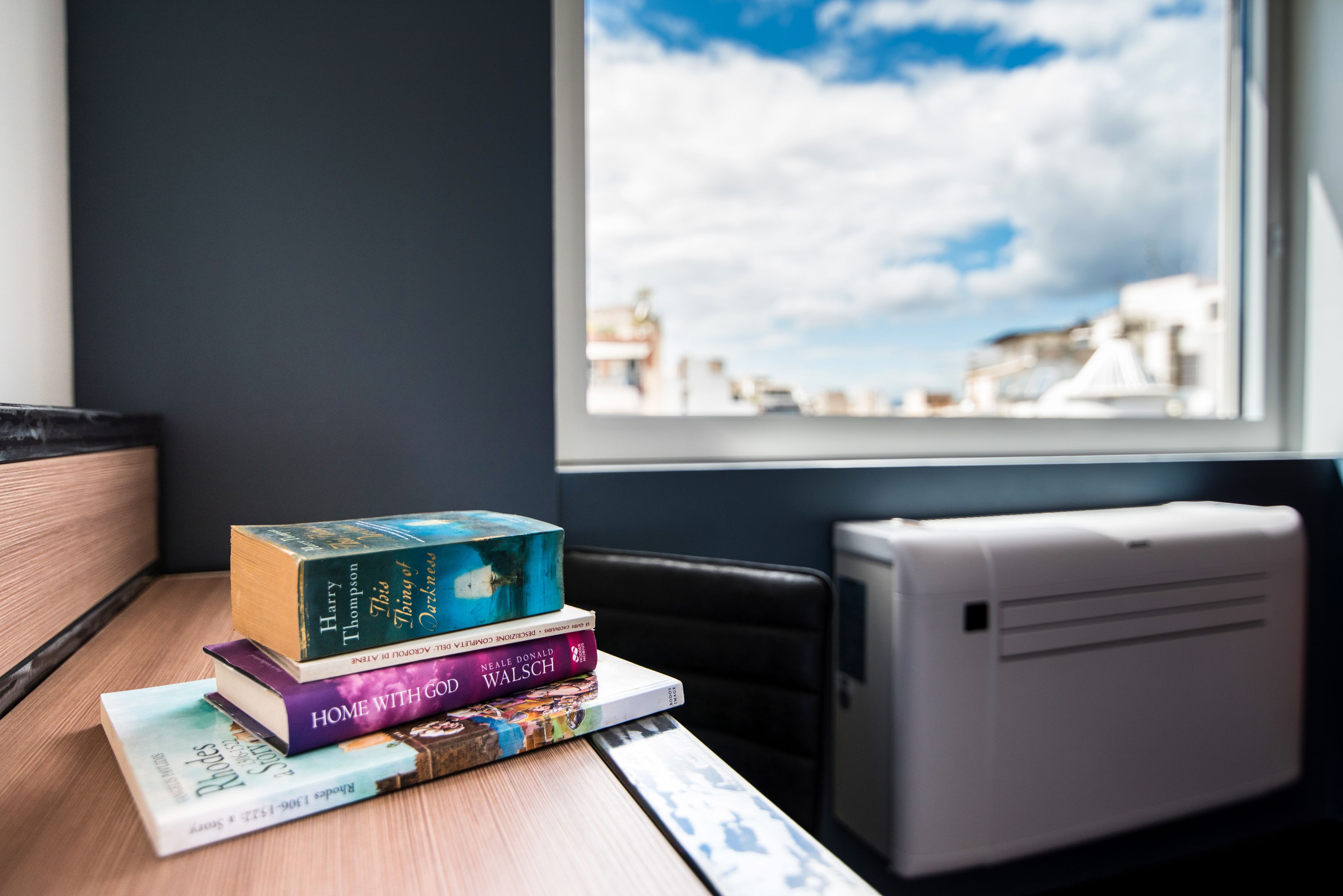 A stack of books on a wooden table by a window with a view of buildings and a blue sky.