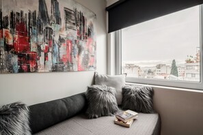 A modern living room with a grey sofa, a large abstract painting, and a view of buildings through a window. - AthinA STREETAPARTMENTS (Athens)