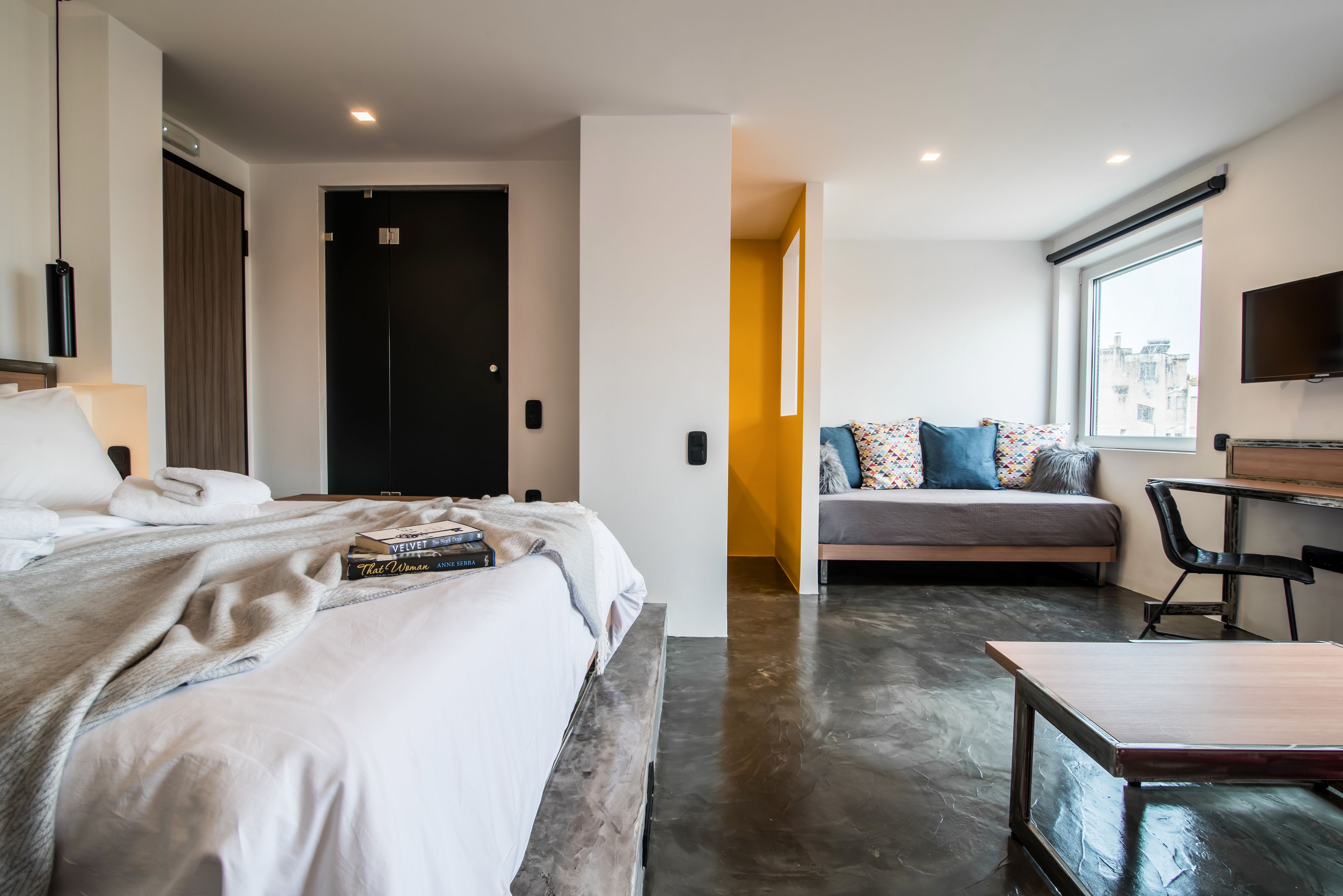 Habitació d'hotel moderna amb un llit gran, una taula de treball i un sofà. Hi ha una porta que dona a una altra habitació i una finestra amb vista a la ciutat.