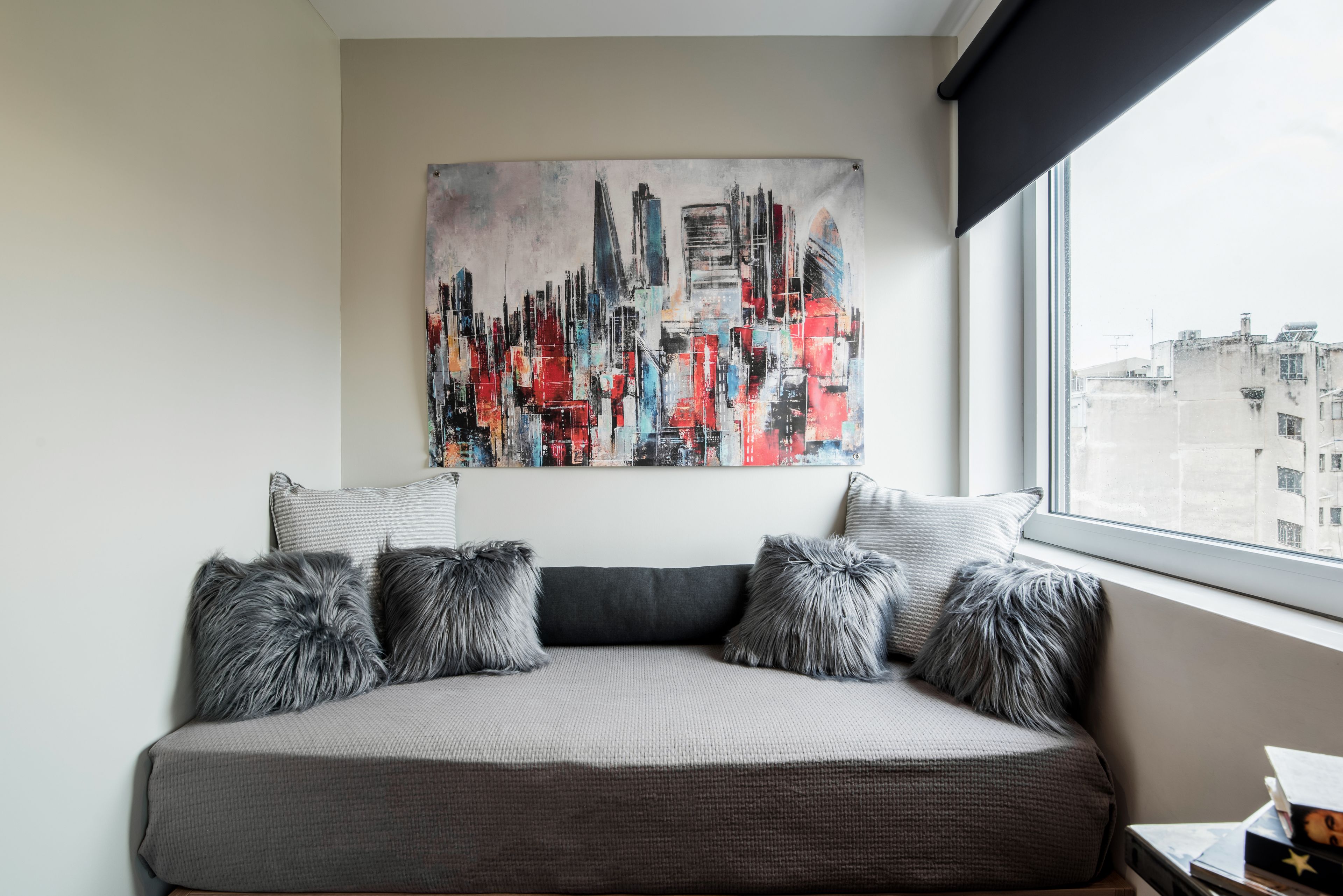 A modern bedroom with a gray sofa bed, fluffy pillows, a cityscape painting, and a window with a view of buildings.