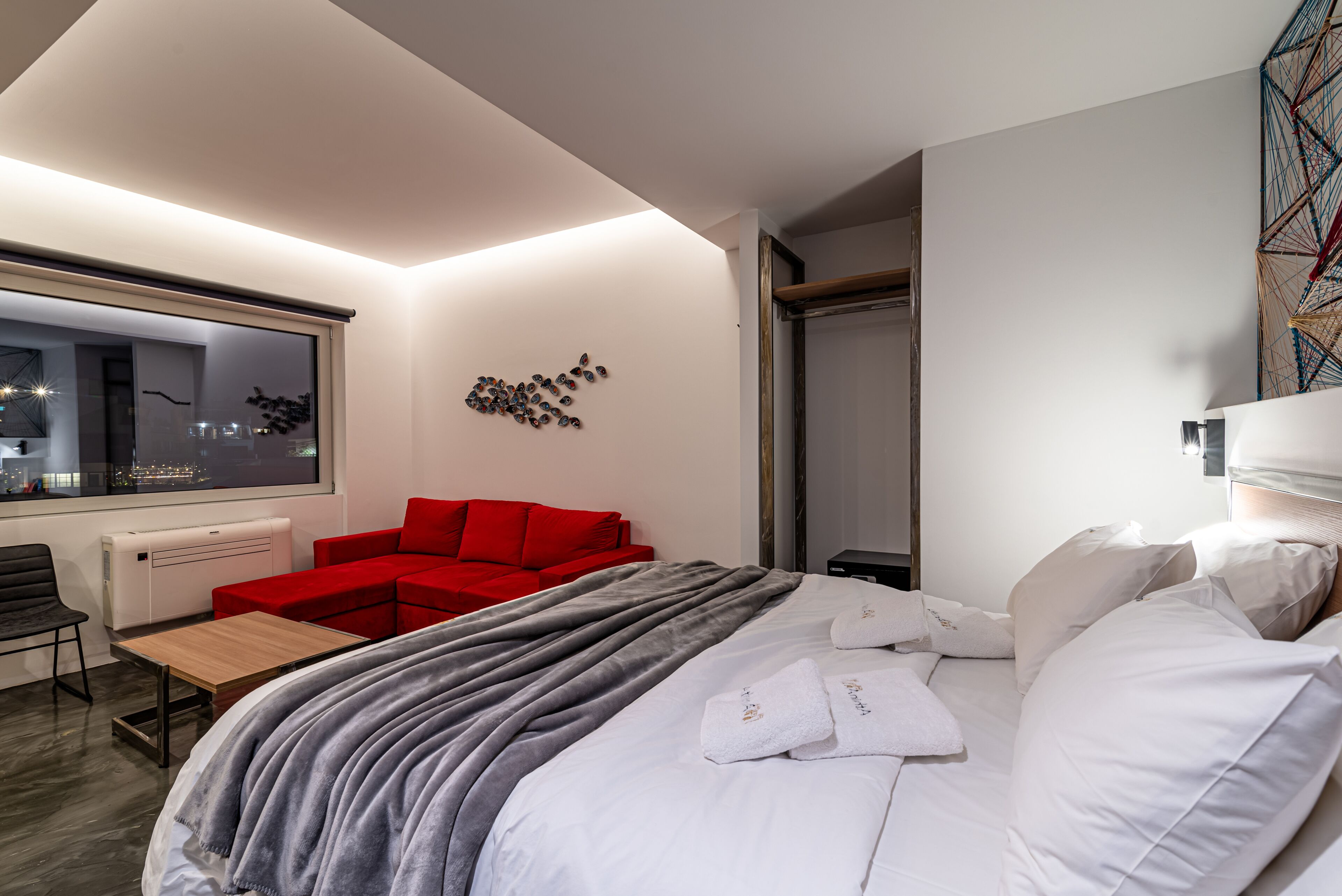 A modern hotel room with a large bed, a red sofa, a wooden table, and a window with a city view.