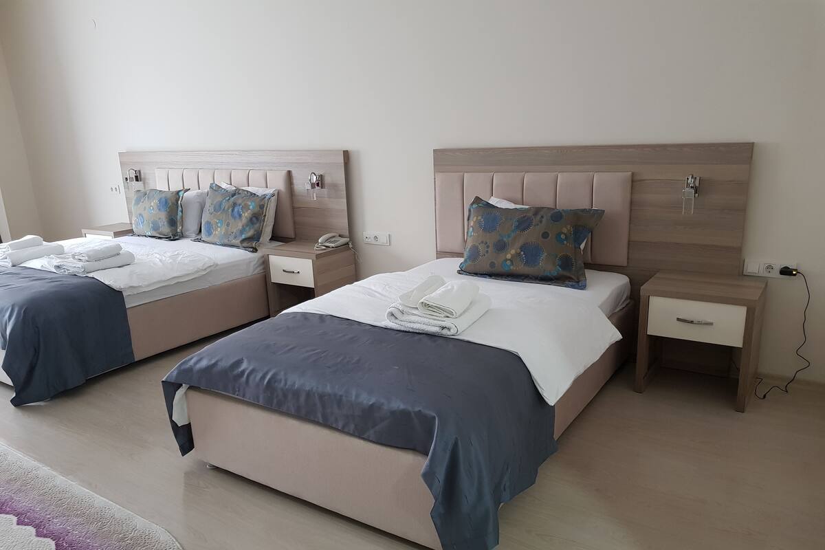 A hotel room with two beds, each with a bedside table, a phone, and a lamp. The room has a wooden headboard and a light-colored floor.