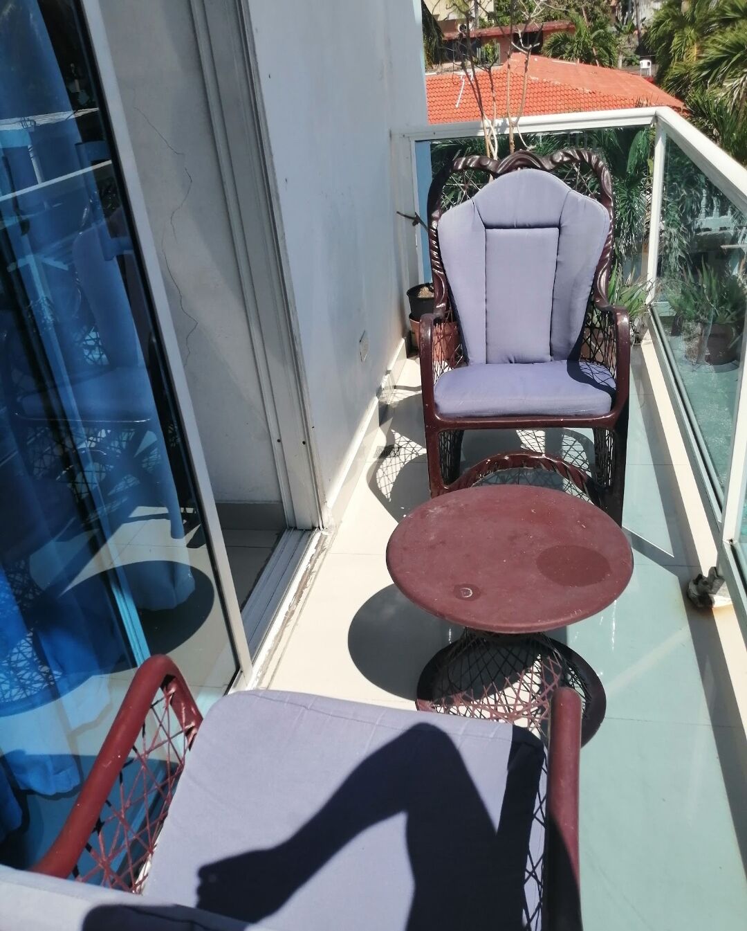 Uma varanda com porta de vidro, uma cadeira, uma mesa pequena e vista para um edifício com telhado vermelho e vegetação.