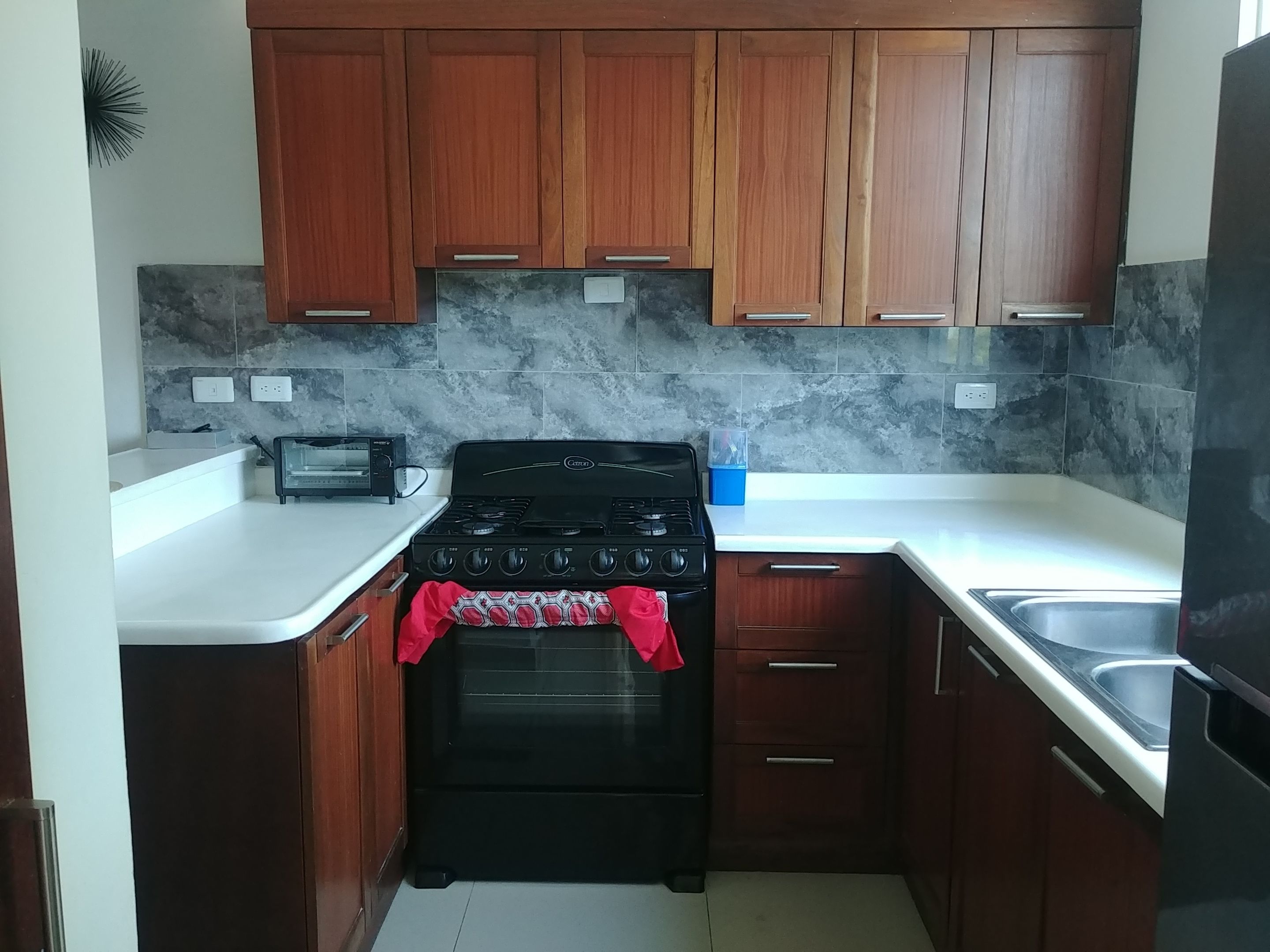 A compact kitchen with wooden cabinets, a black stove, a white countertop, and a tiled backsplash.