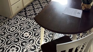 A kitchen with white cabinets, a stainless steel sink, a stove, and a refrigerator. There's a window with blinds, a patterned floor, and a table with a paper on it.