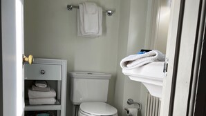 A bathroom with a toilet, a shelf with folded towels, a radiator, and a white rug on the floor.