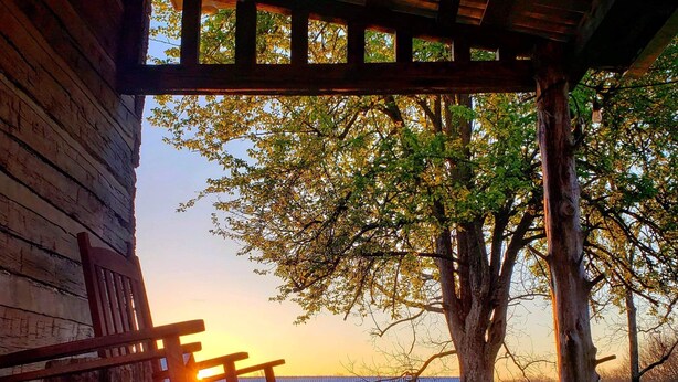A wooden porch with a rocking chair, a view of a tree, and a sunset in the background.
