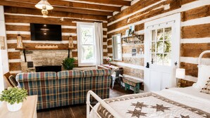 A log cabin interior with a sofa, a bed, a fireplace, a TV, and a window with curtains.