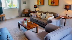 A living room with a sofa, armchairs, a coffee table, and a TV. There are two lamps, a picture on the wall, and a window with curtains. - Rural peace & luxury near Malton, beautiful barn, stunning views, woodburner (Malton)