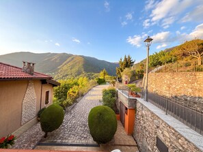 Un chemin pavé mène à un mur de pierre surmonté d’une clôture métallique, le tout étant entouré de verdure et ayant une chaîne de montagnes en arrière-plan.