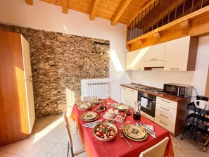 Un espace repas agrémenté d’un mur en pierre, de poutres apparentes en bois au plafond, d’une cuisine équipée et d’une table mise pour le repas.