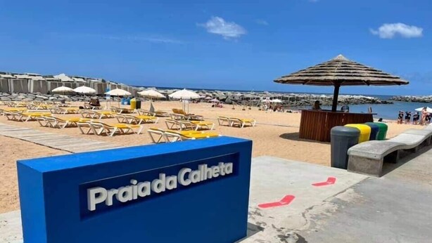 Een bord aan het strand dat Praia da Calheta aankondigt, met ligbedden en parasols op de achtergrond.