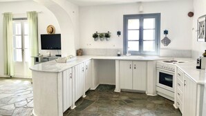A kitchen with white cabinets, a stone floor, a window with blue trim, and a flat-screen TV on the wall.