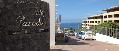 A stone sign with "Residencial Adoje Paradise" written on it, leading to a road with parked cars and a building with a white facade and a balcony.