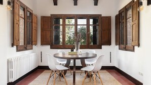 A dining area with a round table, white chairs, wooden cabinets, and a view through large windows. - Spacious Luxury Villa with Heated Pool, A/C, Near Barcelona, Vineyards & Golf (Castellví de Rosanes)