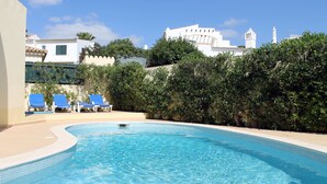 Une piscine aux carreaux bleus, entourée de verdure et bordée de bâtiments blancs en arrière-plan.