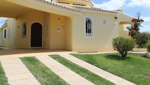 Un bâtiment à deux étages, doté d’une façade jaune, d’une entrée centrale avec une porte et d’un petit jardin agrémenté d’un arbre et d’un pot de fleurs.
