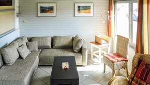 Un salon avec un canapé d’angle gris, une table basse noire et un fauteuil en osier avec un coussin à carreaux. Deux tableaux encadrés sont accrochés au mur, un climatiseur blanc est placé au-dessus du canapé, et une fenêtre avec des rideaux se trouve à droite.