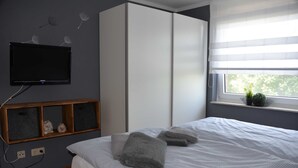 A bedroom with a bed, grey towels, a wardrobe, a TV, and a window with a plant.