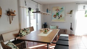A dining area with a long wooden table, wicker chairs, and a bench. There is a painting on the wall and a tall floor lamp.