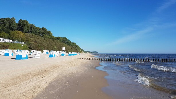 A beach with a row of blue and white beach chairs and umbrellas, a wooden jetty, and a forested hillside in the background.