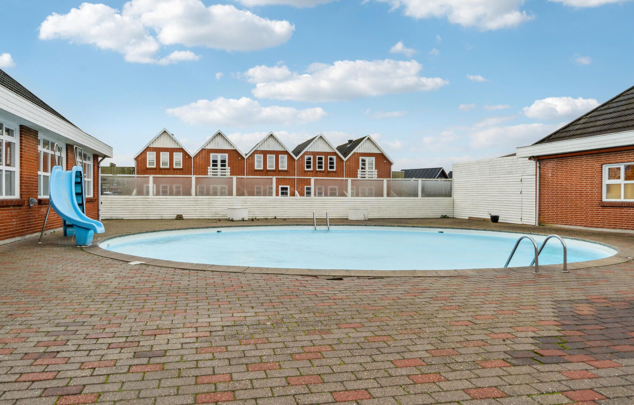 Een zwembad met een blauwe glijbaan, omgeven door een terras van baksteen en een rij rode bakstenen huizen met witte details.