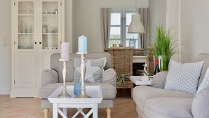 Ein Wohnzimmer mit einer Couch, einem Sessel und einem kleinen Tisch mit Kerzen. Ein Schrank mit Glastüren und ein Fenster mit Vorhängen sind ebenfalls zu sehen.