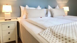 Ein ordentlich bezogenes Bett mit weißen Leinentüchern und einer Decke mit Sternenmuster, das von zwei Nachttischlampen auf einem weißen Nachttisch mit Schubladen flankiert wird.