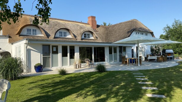 Ein Haus mit Strohdach, einer überdachten Terrasse mit Gartenmöbeln und einem gepflegten Rasen.