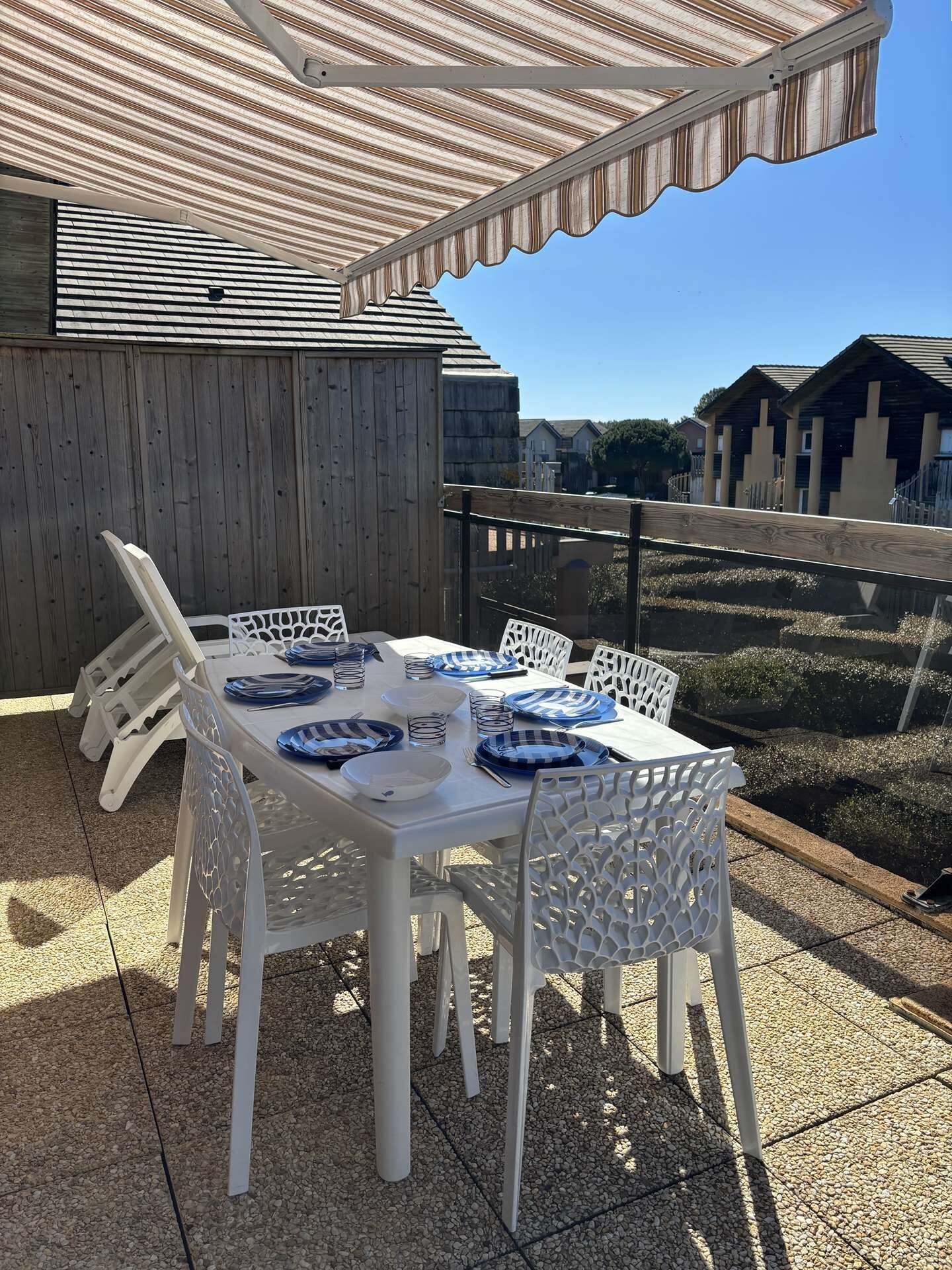 An outdoor patio with a table set for four, surrounded by white chairs, under a striped awning.