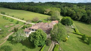 Vue aérienne d’une propriété rurale avec une grande maison entourée d’une végétation luxuriante, d’un jardin bien entretenu et d’un ciel dégagé.