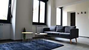 Una sala de estar moderna con un sofá gris, dos mesitas redondas, un tapete azul oscuro y ventanas amplias.