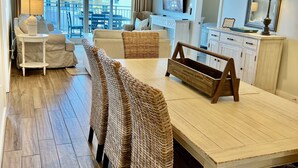 Un comedor con una mesa de madera y sillas de mimbre, una consola blanca con un televisor, y vistas a un edificio a través de amplios ventanales.
