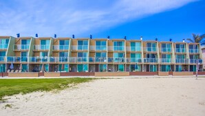 Un hotel frente a la playa con varios balcones, ventanas amplias y una playa arenosa frente a él.