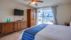 Habitación de hotel con cama, televisor, cómoda de madera y balcón con vista a palmeras.