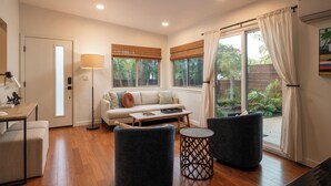 A modern living room with a sofa, two armchairs, a coffee table, and a lamp. There is a door leading to a balcony with a view of greenery.