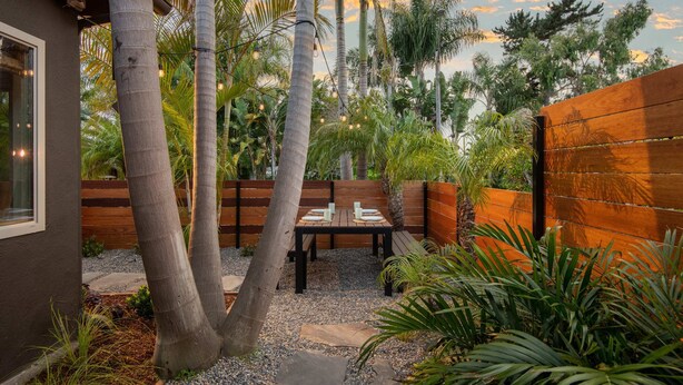 A patio with a wooden table and chairs, surrounded by palm trees and a wooden fence.