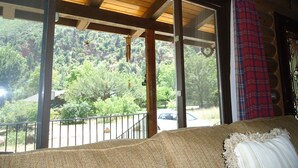 Ein Zimmer mit großem Fenster und Blick auf eine bewaldete Landschaft, Holzboden und einem roten Karomuster-Vorhang.
