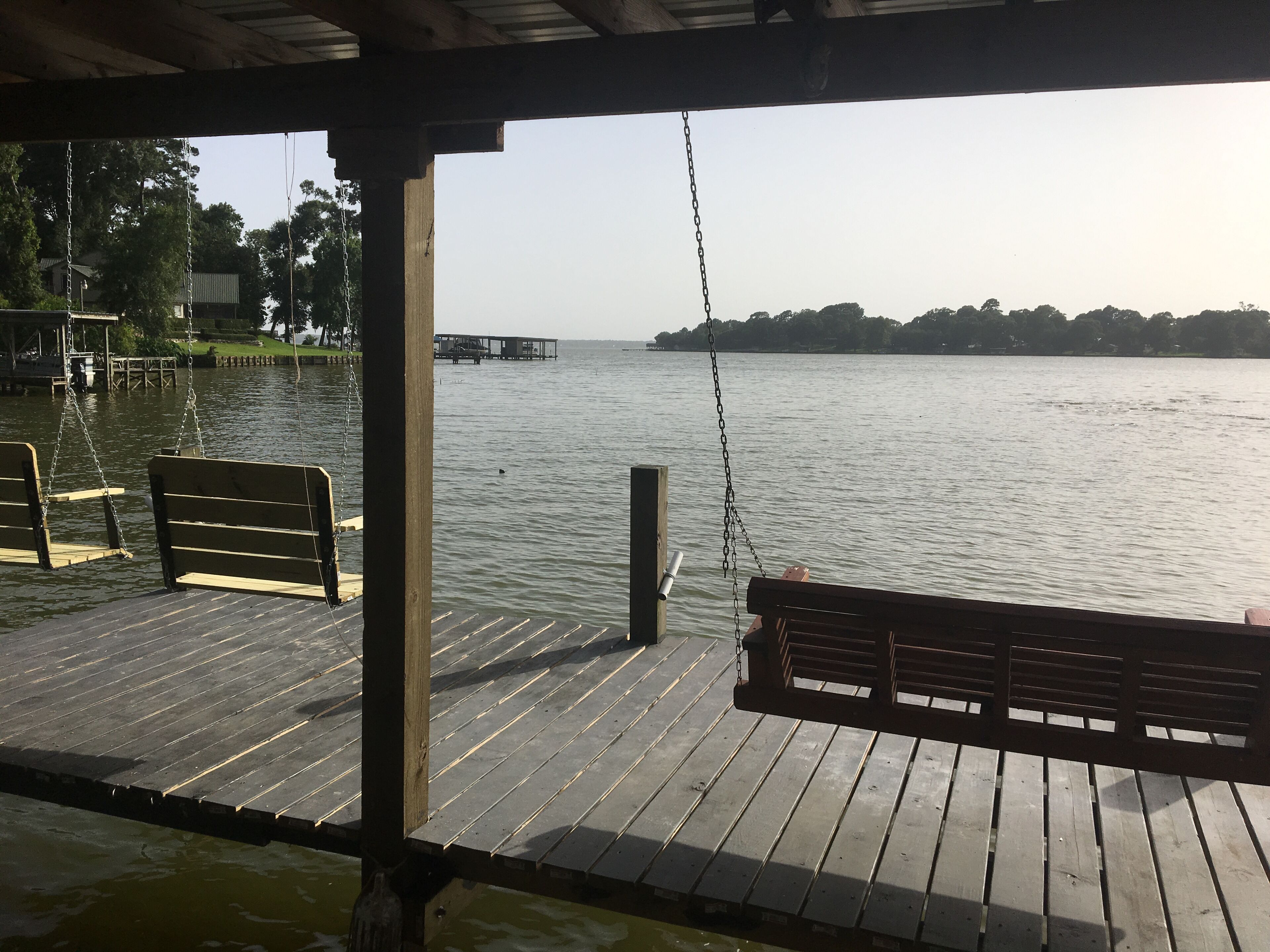 Eine Holzstege mit Bänken, der auf ruhiges Wasser blickt, ein weiter Dock im Hintergrund und Bäume.