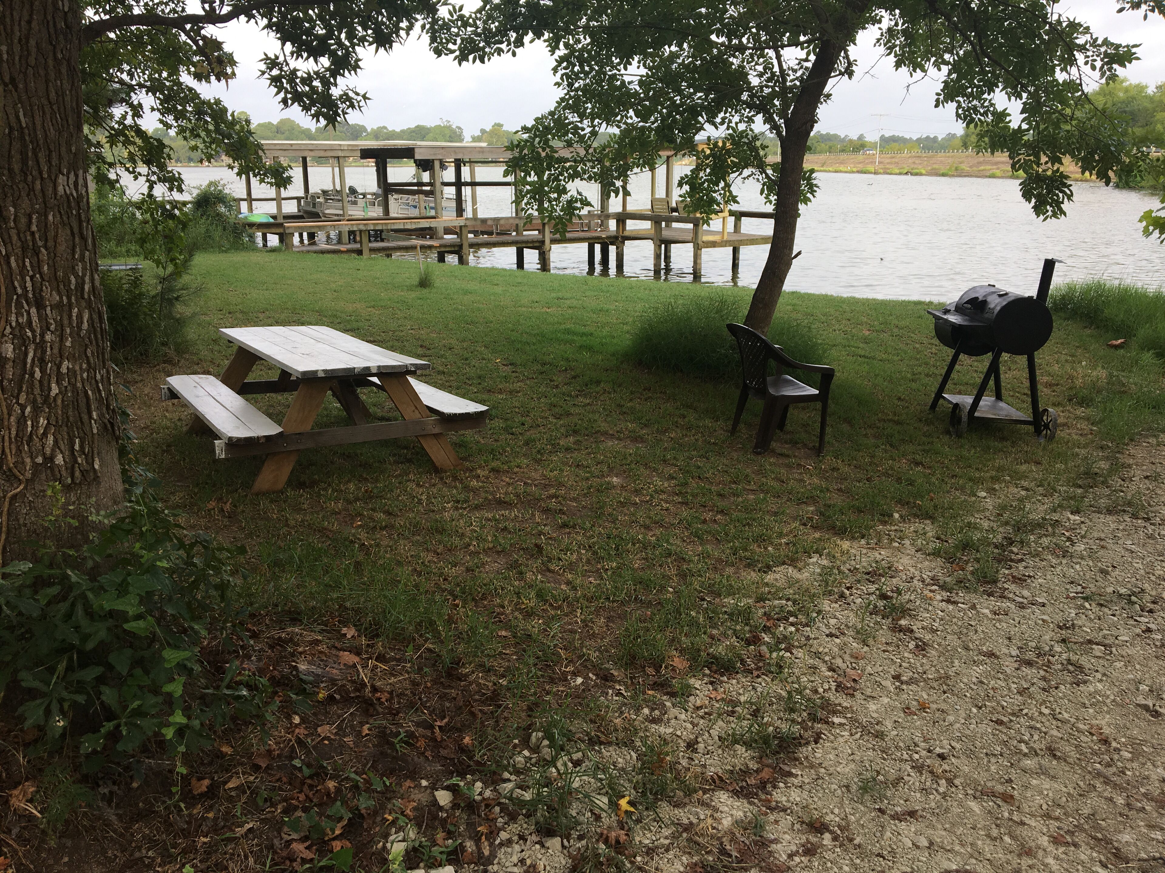 A picnic area with a wooden table and benches, a grill, and a dock extending into a lake.