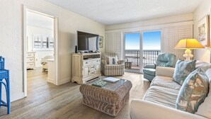 A spacious living room with a large window, a television, a wicker ottoman, and a sofa with cushions.