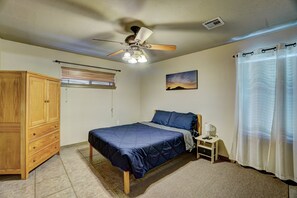 Une chambre à coucher comprenant un lit, une armoire en bois, une table de chevet, un ventilateur de plafond, une fenêtre avec des stores, un tableau et un rideau léger.