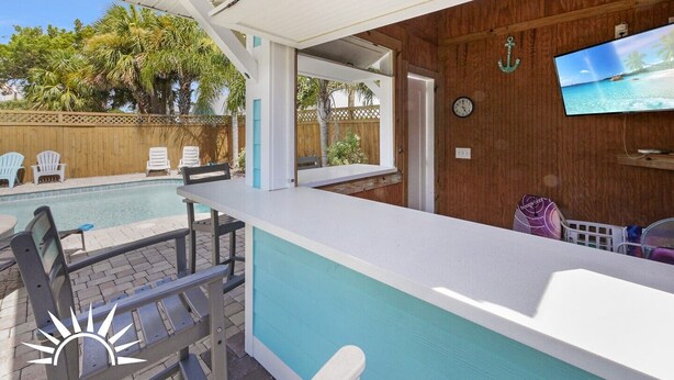 A poolside area with a covered seating area, a television mounted on the wall, and a wooden fence in the background. - Sapphire: Updated 2 bedroom home with private pool, hot tub, cabana, outdoor (St. Augustine Beach)
