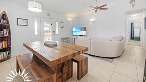 A spacious living area with a wooden dining table, a white sofa, a television, and a ceiling fan. - Sapphire: Updated 2 bedroom home with private pool, hot tub, cabana, outdoor (St. Augustine Beach)