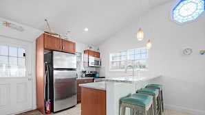 A kitchen with wooden cabinets, stainless steel appliances, a central island with bar stools, and a view of the outside through large windows. - Sapphire: Updated 2 bedroom home with private pool, hot tub, cabana, outdoor (St. Augustine Beach)