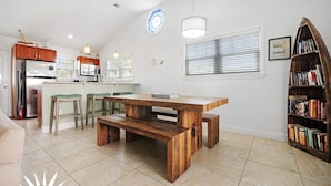 A modern kitchen with a central island, a dining area with a wooden table and bench, and a bookshelf filled with books. - Sapphire: Updated 2 bedroom home with private pool, hot tub, cabana, outdoor (St. Augustine Beach)