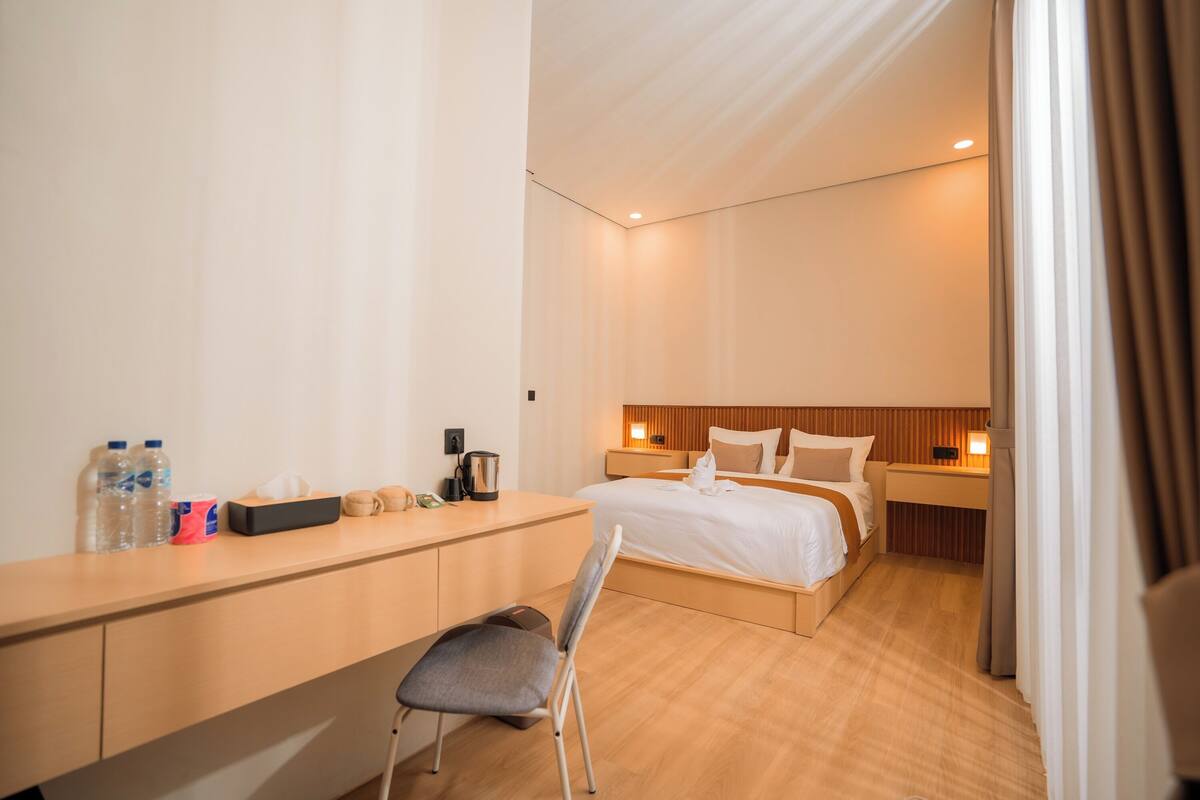 A modern hotel room with a wooden desk, a bed with white linens, and a nightstand with a lamp.