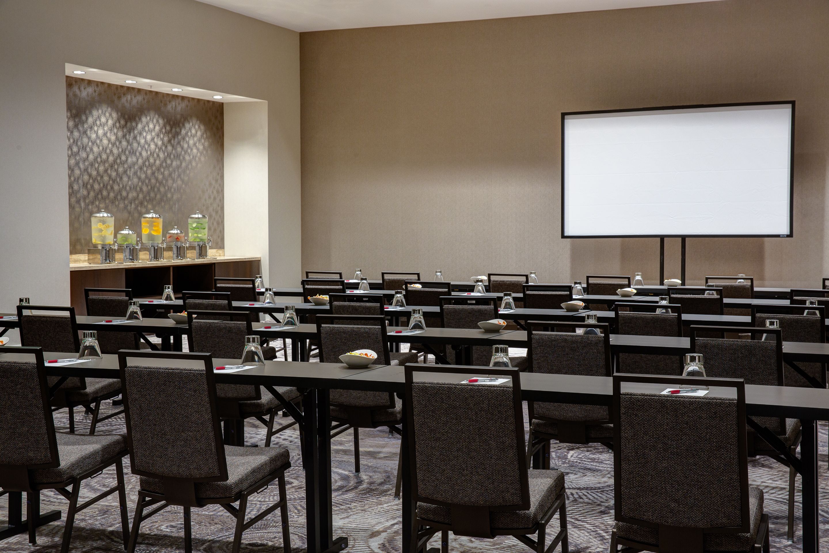 A conference room with rows of tables, chairs, a projector screen, and a beverage station.